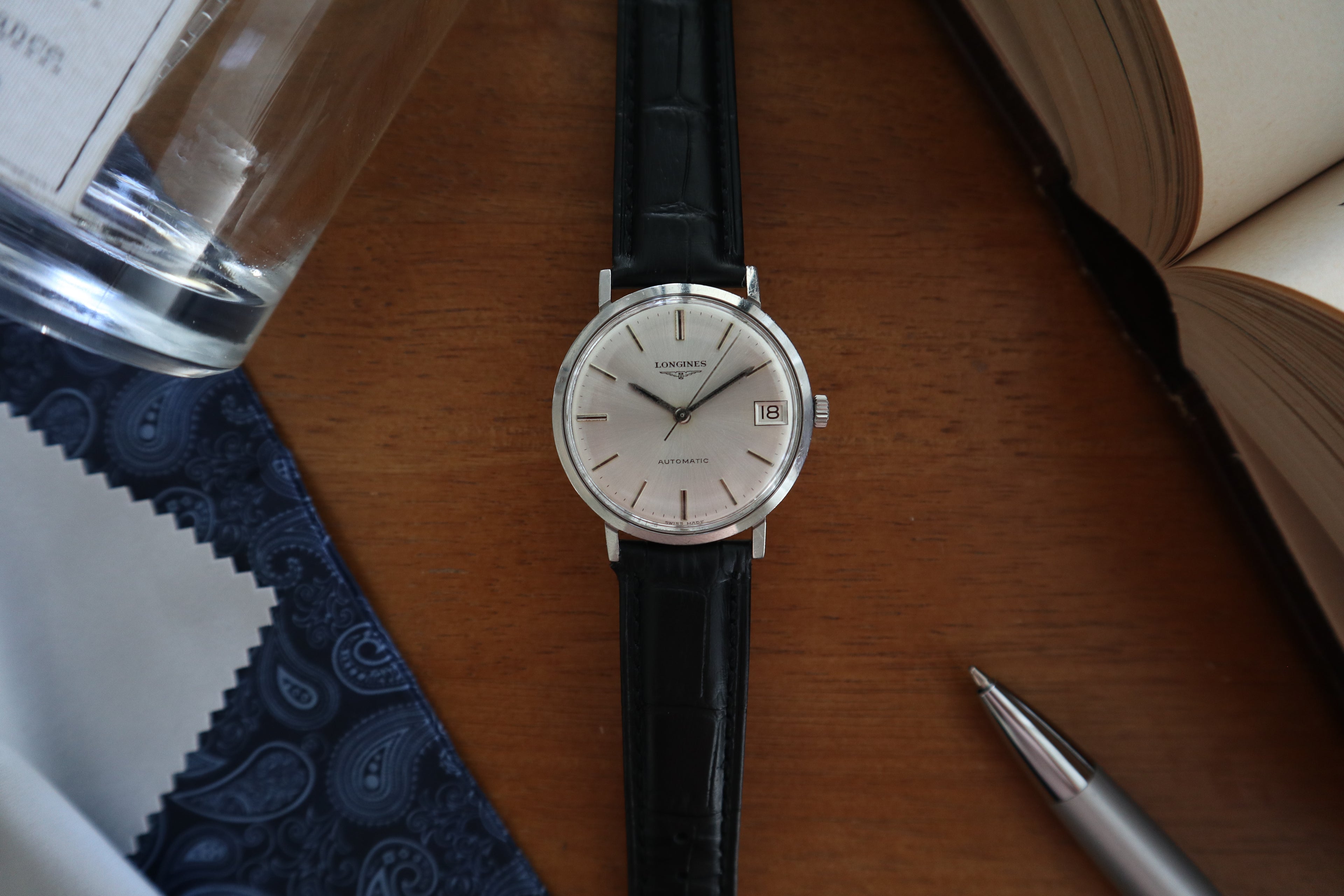 Vintage Longines Automatic Date watch with silver dial and black leather strap resting on a wooden desk beside a glass of water and open book.