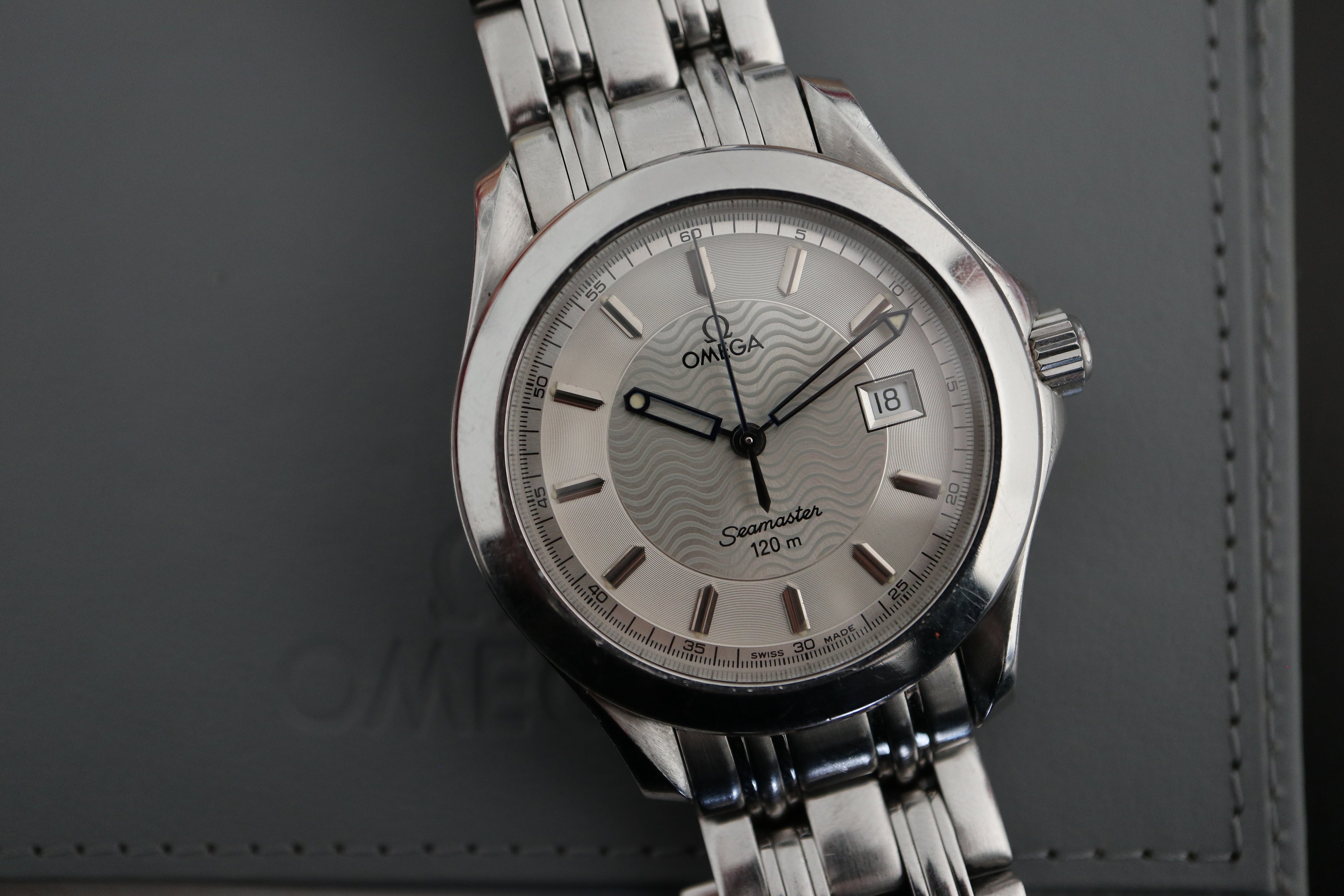Omega Seamaster 120M Pre-Bond with silver wave dial and stainless-steel bracelet resting on a grey Omega pouch.