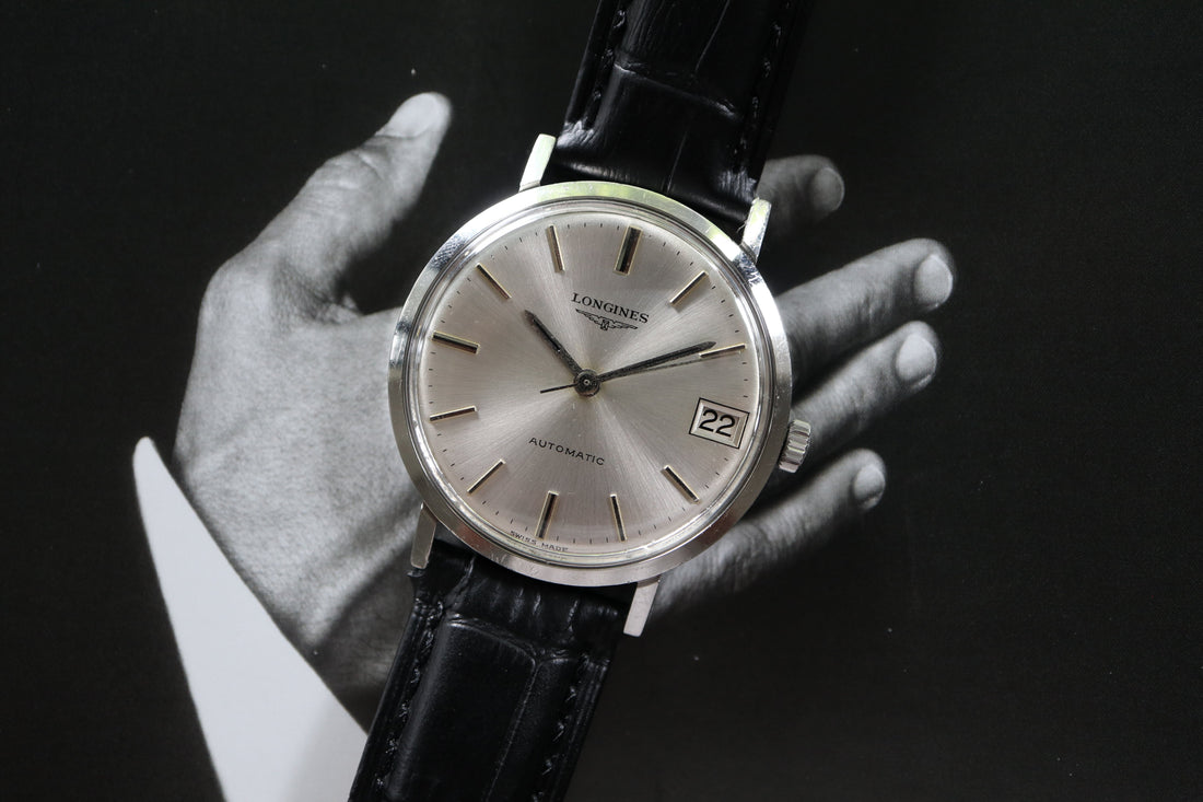 A vintage Longines automatic wristwatch with a silver sunburst dial, slim baton hour markers, and a date window at 3 o’clock. The watch is fitted on a black leather strap and photographed against a monochrome background showing a man’s hand in a suit.